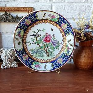 Op de afbeelding: Een decoratieve porseleinen bord met een bloemen- en vogelontwerp. De plaat heeft een centraal beeld van een vogel die op een bloeiende tak zit, omgeven door een blauwe en gouden rand. De plaat wordt op een gouden standaard gepresenteerd.
