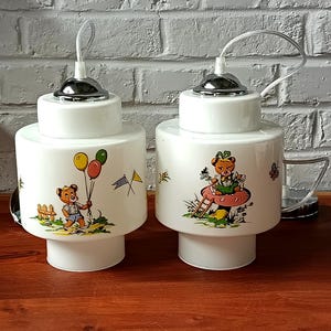 Puede incluir: Dos lámparas colgantes de cerámica blanca con detalles cromados. Cada lámpara presenta un cuerpo cilíndrico con una ilustración de oso de dibujos animados sosteniendo globos y sentado en una seta. Ideal para una habitación infantil.