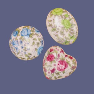 Può includere: Tre elementi decorativi in porcellana con motivi floreali. Uno è rotondo con fiori blu, un altro ovale con fiori verdi e il terzo a forma di cuore con rose rosa. Tutti hanno una base bianca e sono su uno sfondo blu.
