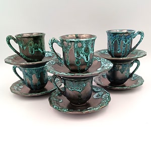 Op de afbeelding: Zes keramische espressokopjes en schotels. De kopjes en schotels hebben een donkerbruine basis met een turquoise en groene glazuur die langs de zijkanten druipt. De schotels hebben een geschulpte rand. De kopjes hebben een gebogen handvat.