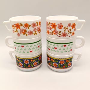 Puede incluir: Dos pilas de tres tazas de cerámica blanca. Cada taza presenta un patrón floral diferente en naranja, rosa, verde y azul. La capa superior tiene flores naranjas y rosas, la capa media tiene hojas verdes y bayas rojas, y la capa inferior tiene flores azules.