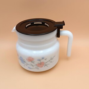 以下が含まれることがあります： 黒い蓋とハンドルが付いた白いガラスピッチャー。ピッチャーは、青とサンゴの色合いの花柄のデザインが特徴です。ピッチャーは飲み物を提供するように設計されています。