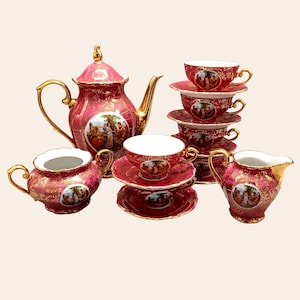 Puede incluir: Un juego de té vintage en tonos rojos y dorados. El juego incluye una tetera, azucarero, lechera y varias tazas y platillos. Cada pieza presenta un ribete dorado y una escena decorativa.