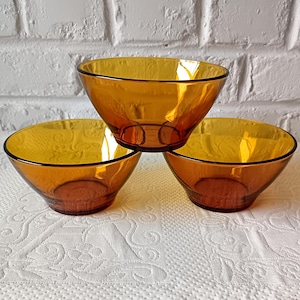 May include: Three amber glass bowls are arranged on a white patterned tablecloth. The bowls have a rounded shape with a slightly flared rim and a dark rim. The bowls are made of transparent glass.