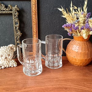 May include: Two clear glass beer mugs with handles. The mugs have a decorative base and are sitting on a wooden surface. A decorative vase with dried flowers is in the background.