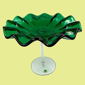 May include: A green glass serving dish with a wavy edge and a clear glass stem. The dish is on a white background.