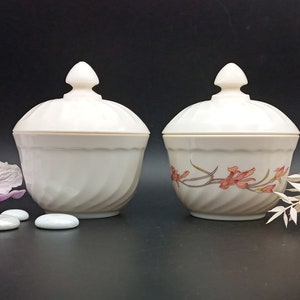 Peut inclure: Deux bols en céramique blanche avec couvercle et décor floral. Un bol a un couvercle blanc uni et l'autre a un décor floral sur le couvercle. Les bols sont sur une surface noire.