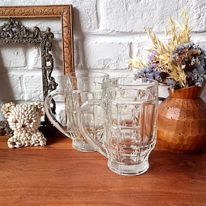 Op de afbeelding: Twee heldere glazen bierpullen met handvatten, met een rasterpatroon, staan op een houten oppervlak. Een decoratief houten frame en een kleine sculptuur staan op de achtergrond. Een vaas met gedroogde bloemen is ook zichtbaar.