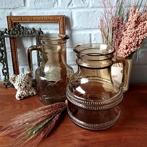 Peut inclure: Deux carafes en verre ambré avec des poignées. L'une présente un motif texturé, l'autre un détail en forme de corde au milieu. Les carafes sont posées sur une surface en bois, avec des éléments décoratifs en arrière-plan.