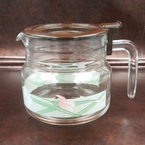 Puede incluir: Una jarra de café de vidrio transparente con tapa y asa de plástico marrón. La jarra tiene un diseño floral en rosa, blanco y verde. La jarra está diseñada para contener líquido y tiene un pico vertedor.