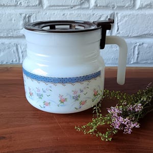 May include: White vintage pitcher with a brown lid and handle. The pitcher features a blue patterned band with floral designs in pink, purple, and green. The handle is white, and the pitcher sits on a wooden surface.
