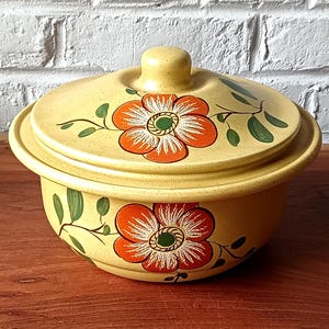 Peut inclure: Un plat à casserole en céramique jaune avec un couvercle. Le plat et le couvercle sont décorés de fleurs orange et de feuilles vertes. Le plat à casserole est posé sur une surface en bois.
