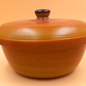 Könnte beinhalten: Eine runde Servierschüssel mit Deckel in Braun- und Orangetönen. Die Schüssel hat einen breiten Rand und einen passenden Deckel mit einem kleinen, dunkelbraunen Knauf. Die Oberfläche der Schüssel hat ein subtiles, strukturiertes Aussehen, das auf Keramik oder Steinzeug hindeutet.