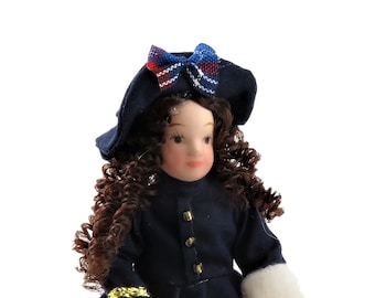 Puede incluir: Una muñeca de porcelana que lleva una falda de cuadros azules y rojos, una chaqueta azul marino con ribetes dorados y un sombrero negro con un lazo de cuadros. La muñeca tiene el pelo rizado marrón y una cara pintada.