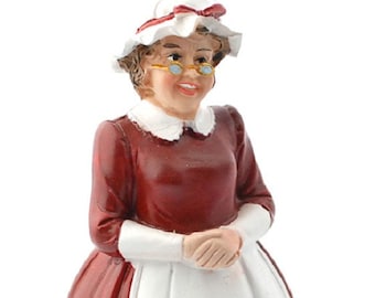 May include: A figurine of a woman wearing a red dress with a white apron and a white hat with a red ribbon. She is wearing glasses and has a friendly expression.