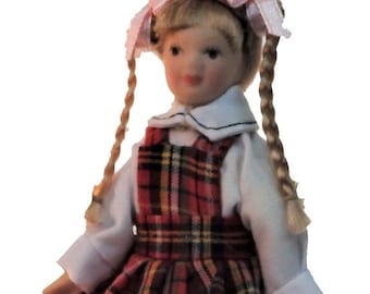 Puede incluir: Una muñeca de porcelana que lleva un vestido de cuadros rojos y negros con una blusa blanca y trenzas. La muñeca tiene el pelo rubio y los ojos azules.