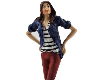May include: A figurine of a woman wearing a blue denim jacket, a white and black striped shirt, and red pants. She is standing with her hands on her hips and looking up.