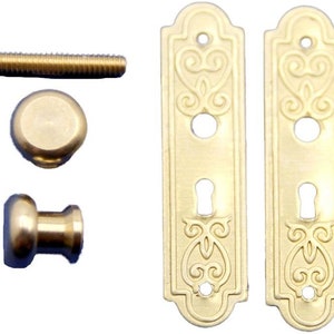 Könnte beinhalten: Ein Satz aus zwei goldfarbenen Türbeschlagplatten mit verzierten Designs und einem Schlüsselloch, sowie zwei Messingknöpfen und einer Schraube.