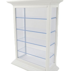 Puede incluir: Vitrina de madera blanca con cuatro estantes de vidrio. La vitrina tiene un borde decorativo alrededor de la parte superior e inferior.