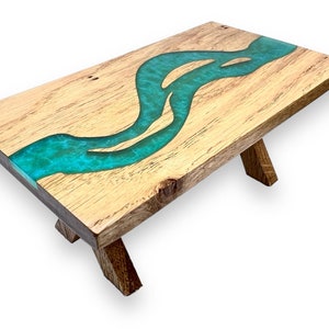May include: A wooden coffee table with a turquoise blue resin river flowing through the center. The table has a natural wood finish and is supported by two wooden legs.