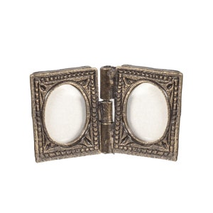 Puede incluir: Un marco de fotos de metal dorado con dos aberturas ovaladas. El marco está articulado en el medio y tiene un diseño ornamentado.