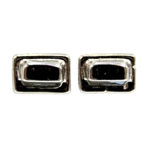 Puede incluir: Dos pendientes de botón rectangulares plateados. Cada pendiente presenta un centro rectangular en relieve con un interior oscuro, posiblemente negro. Los pendientes tienen un diseño geométrico simple con una apariencia ligeramente texturizada.