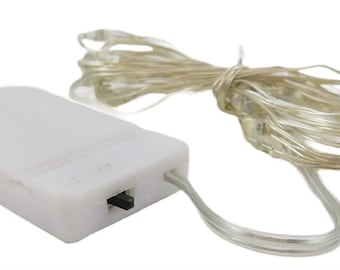 Puede incluir: Un paquete de baterías blanco con una cadena de alambre transparente de luces LED blancas. El paquete de baterías tiene un interruptor en el lateral.