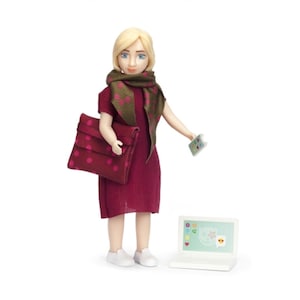 May include: A doll wearing a burgundy dress, a green and pink scarf, and holding a red polka dot purse. The doll is standing next to a white laptop with a colorful screen.