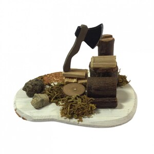 May include: A miniature wooden scene depicting a woodcutter's tools. A small wooden axe is propped against a stack of logs, with a small pile of wood chips and moss surrounding the base. The scene is set on a white wooden base.