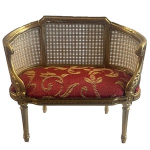 May include: Miniature gold-colored sofa with a red cushion featuring a gold floral pattern. The back and sides have a woven cane design. The sofa has four delicate legs, and the overall style is ornate and elegant.