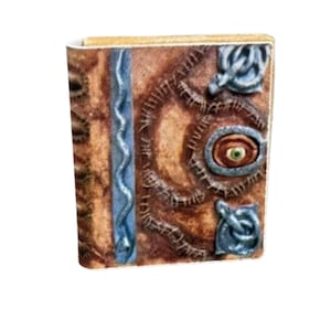 Puede incluir: Un diario de cuero marrón con un diseño de serpiente azul, un ojo cosido con un iris verde y un cierre de metal azul.