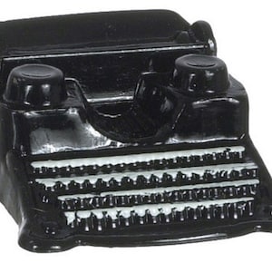Puede incluir: Una figurita de máquina de escribir vintage negra con un teclado blanco. La máquina de escribir tiene una forma redondeada y dos botones negros en la parte superior.