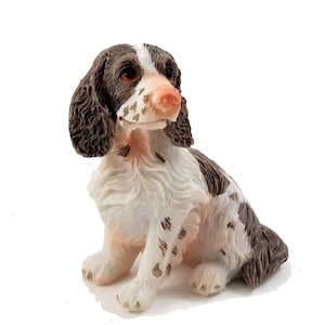 May include: A brown and white figurine of a sitting Springer Spaniel dog. The dog has brown ears, a white body with brown spots, and a brown tail.