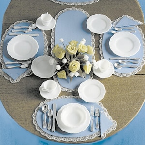 May include: A table setting with white plates, blue placemats, and silver cutlery. A bouquet of yellow roses is in the centre of the table.