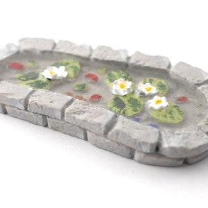 May include: Miniature stone-edged pond with water lilies, lily pads, and small fish. The pond has a gray stone border and a small green frog figurine on the edge. The pond is a decorative item.