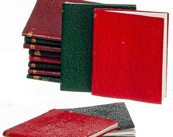 Op de afbeelding: Een verzameling miniatuurboeken met gestructureerde covers in tinten rood, groen en zwart. De stapel heeft goudkleurige accenten op de ruggen, terwijl twee losse boeken plat op de voorgrond liggen en witte pagina's tonen.