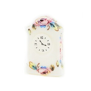 May include: A white porcelain clock with a floral design. The clock face has Roman numerals and black hands. The clock is shaped like a small house.