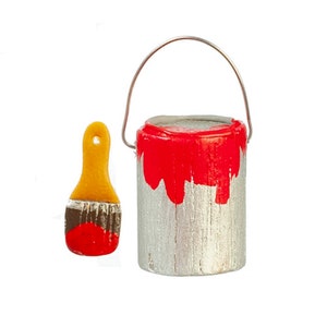 Puede incluir: Un pequeño bote de pintura rojo con una tapa de metal plateado y un asa de alambre. El bote está parcialmente lleno de pintura roja y tiene un pequeño pincel con un mango amarillo a su lado.
