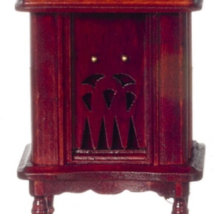 Puede incluir: Un armario de madera de color rojo oscuro con un diseño de corte decorativo en la puerta delantera. El armario tiene un pequeño estante superior y se asienta sobre cuatro patas torneadas.