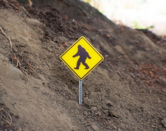 May include: A yellow warning sign with a black silhouette of a Bigfoot walking. The sign is posted on a dirt hill.