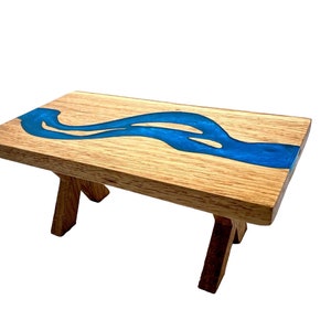 May include: A small wooden table with a blue epoxy river design. The table has a rectangular top and angled legs. The wood has a natural grain, and the blue epoxy flows across the surface, creating a unique decorative piece.