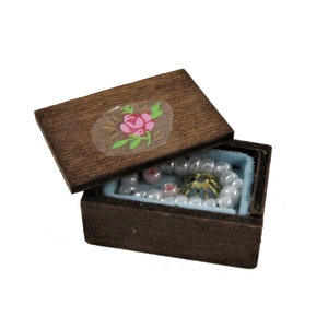 Puede incluir: Una caja de madera con un diseño de rosa pintado en la tapa. La caja está abierta y contiene un collar de perlas y un anillo de oro.