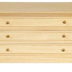 May include: A miniature wooden dresser with three drawers and gold-colored knobs. The dresser is unfinished, with a natural wood grain, and sits on short, angled legs. The top of the dresser has a slight overhang.