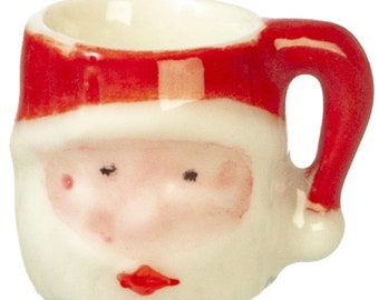 Puede incluir: Una taza de cerámica blanca con una cara de Papá Noel roja y un asa roja. La taza tiene un borde blanco y un interior blanco.