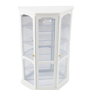 Puede incluir: Vitrina de esquina blanca con puertas y estantes de vidrio. La vitrina tiene una base hexagonal y pomos dorados.