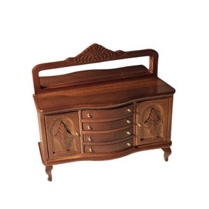 Könnte beinhalten: Miniatur-Buffet aus Holz mit dunkelbraunem Finish. Das Buffet hat vier Schubladen mit goldfarbenen Knöpfen, zwei Türen mit geschnitzten Blumenmustern und eine dekorative Oberseite. Die Beine sind geschwungen.
