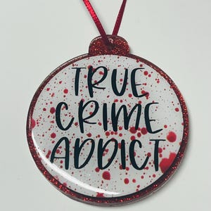 May include: A round ornament with a red glitter border and a red ribbon for hanging. The ornament has a white background with red splatters and the words "TRUE CRIME ADDICT" in black lettering.