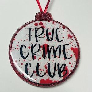 May include: A round ornament with a red glitter border and a red ribbon for hanging. The ornament has the words "TRUE CRIME CLUB" in black lettering, with a white background splattered with red paint.
