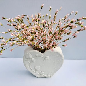 May include: A white heart-shaped vase filled with pink flowers. The vase has a textured surface and a decorative leaf and vine design. The flowers have delicate pink petals and green stems.