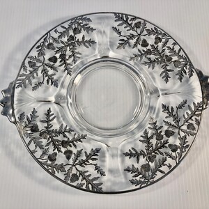 Puede incluir: Plato de servir de vidrio transparente con borde plateado y un patrón decorativo de hojas y bayas. El plato tiene un área circular central y tres secciones divididas. Dos asas plateadas están en lados opuestos.